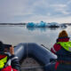 2 persons on a boat, taking a picture in front of blue iceberg.