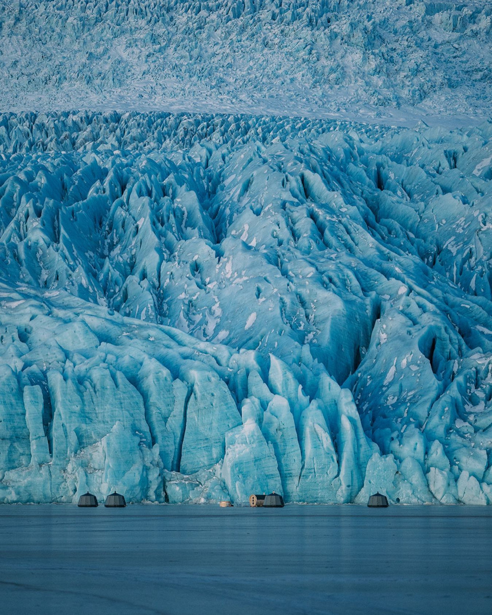 Igloo huts and sauna hut in on a frozen glacial lagoon, in front of a glacier wall.