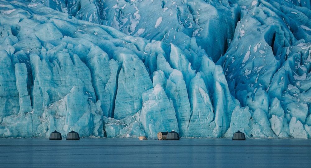 Igloo huts on frozen glacial lagoon, in front of glacier wall.