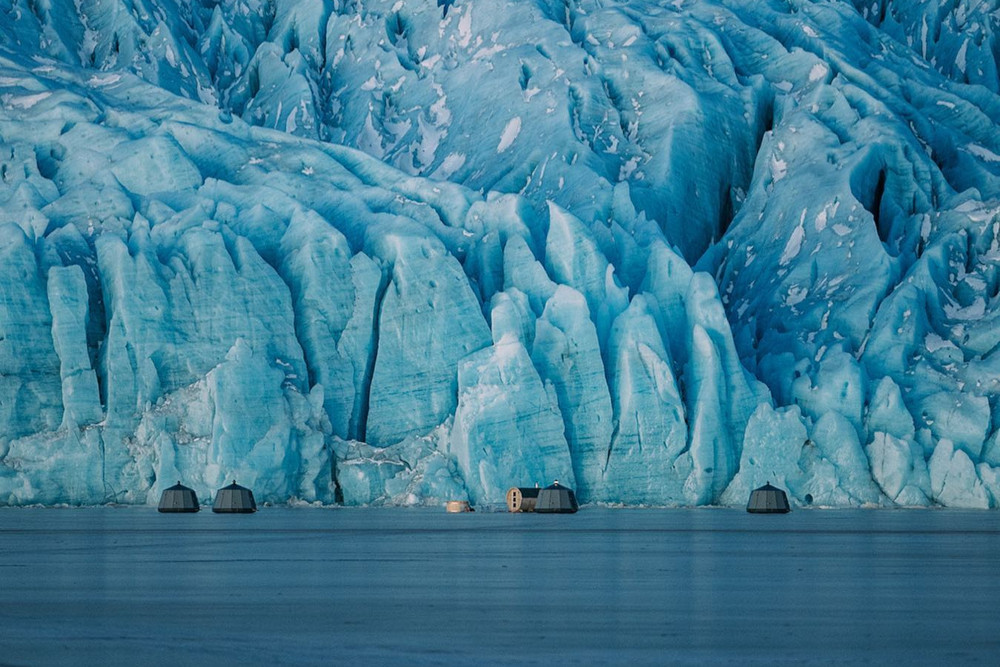 Igloo huts on frozen glacial lagoon, in front of glacier wall.