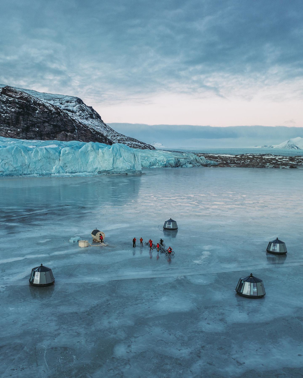 9 persons inside a circle of igloo huts + sauna hut on a frozen glacial lagoon, view from above with drone picture shot. Glacier edge and mountain in background.