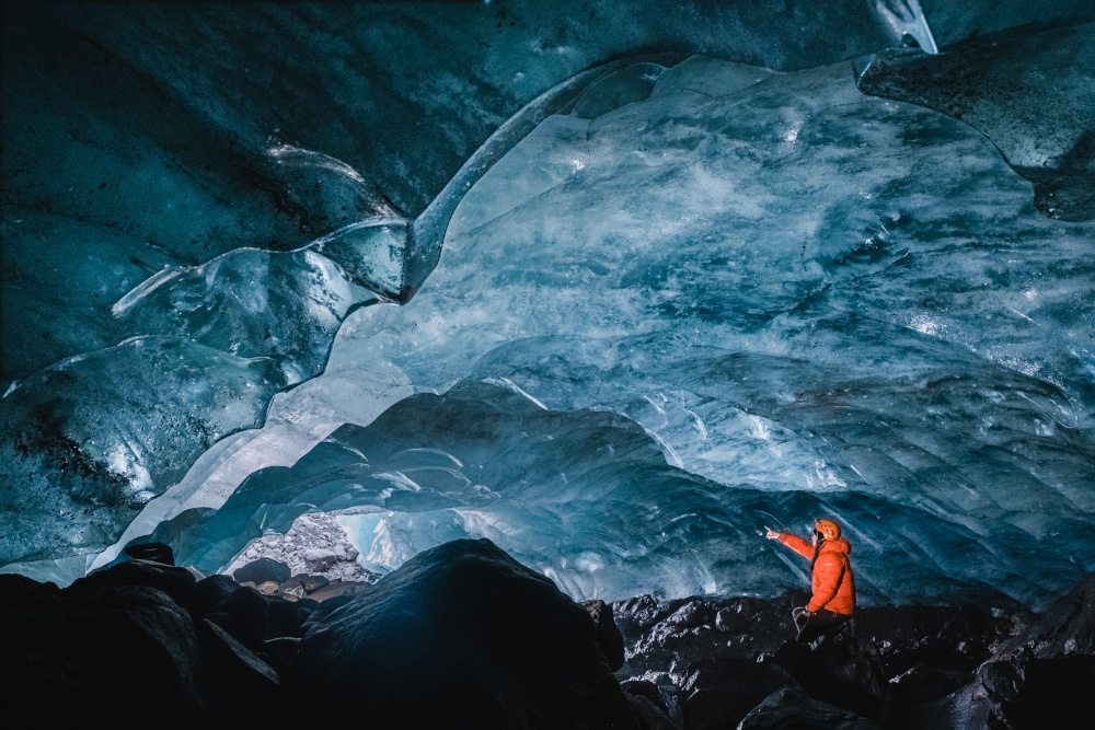 Person in orange jacket under a blue ice cave, pointing to some direction.