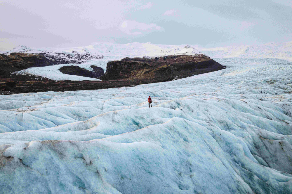 Man standing on hard glacier ice in some distance turning to the mountains further in the background.