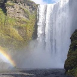 Large waterfall (called Skogafoss) with rainbow and grassy rock wall. Person on picture look really small.