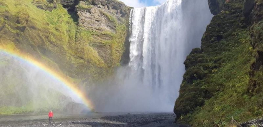 Large waterfall (called Skogafoss) with rainbow and grassy rock wall. Person on picture look really small.