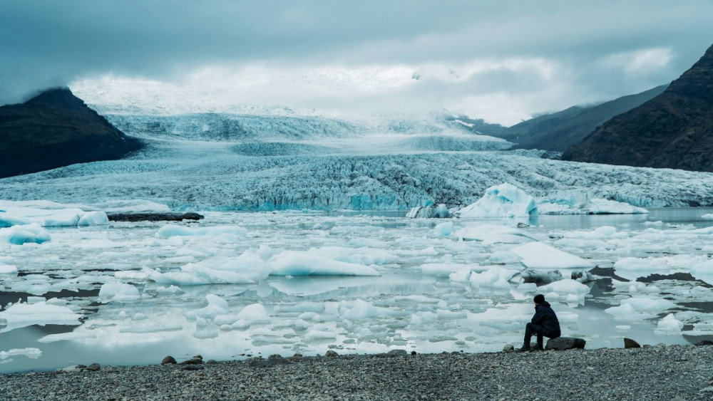 A person sits by the shore at Fjallsárlón glacial lagoon, with many icebergs floating around in the lagoon.