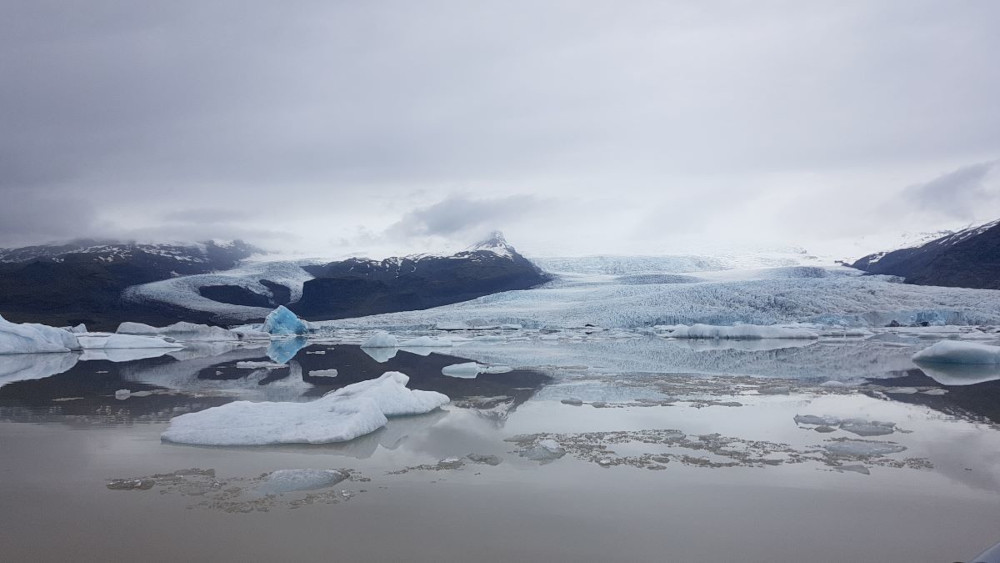 Mirror smooth Fjallsarlón glacial lagoon, with floating icebergs and one blue iceberg in distance and Fjallsjökull glacier in background between two mountains.