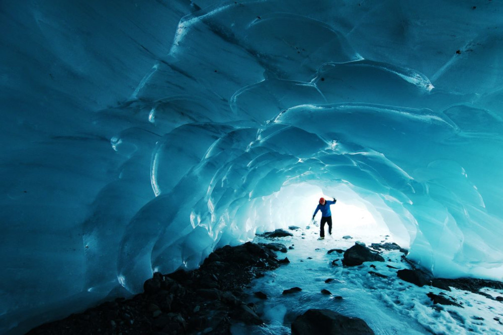 Blue ice tunnel with a person standing in the opening, and the daylight is shining behind the person.