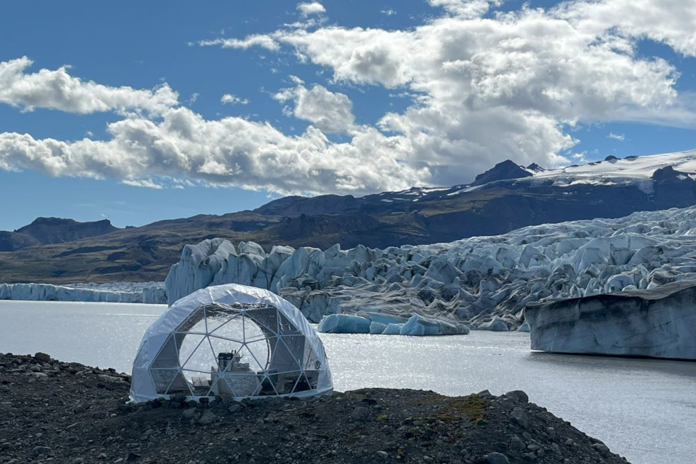 White bubble like tent with windows in front of a glacial lagoon and  glacier wall.