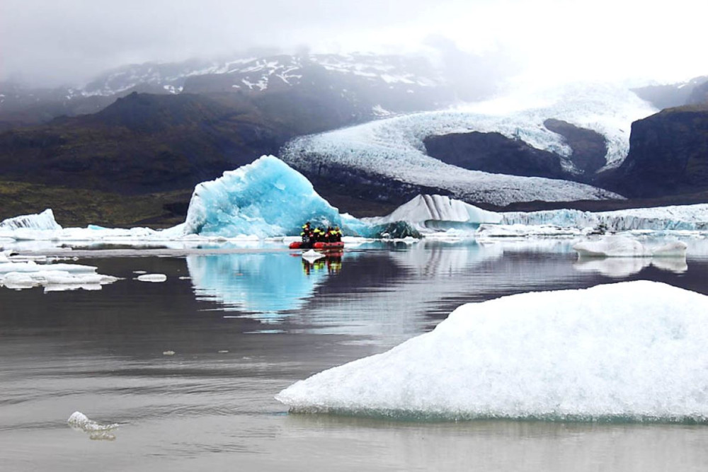 Inflatable boat sailing on Fjallsarlón glacial lagoon with one blue iceberg floating behind in background.