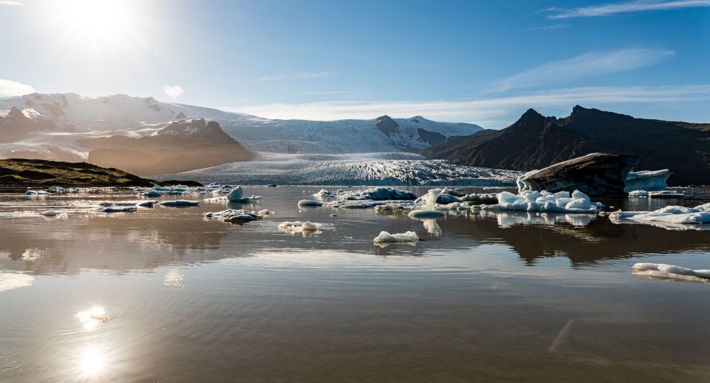 Mirror smooth Fjallsarlon glacier lagoon with the sun in the blue sky, over the glacier mountains.