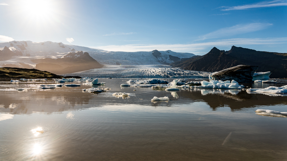 Mirror smooth Fjallsarlon glacier lagoon with the sun in the blue sky, over the glacier mountains.