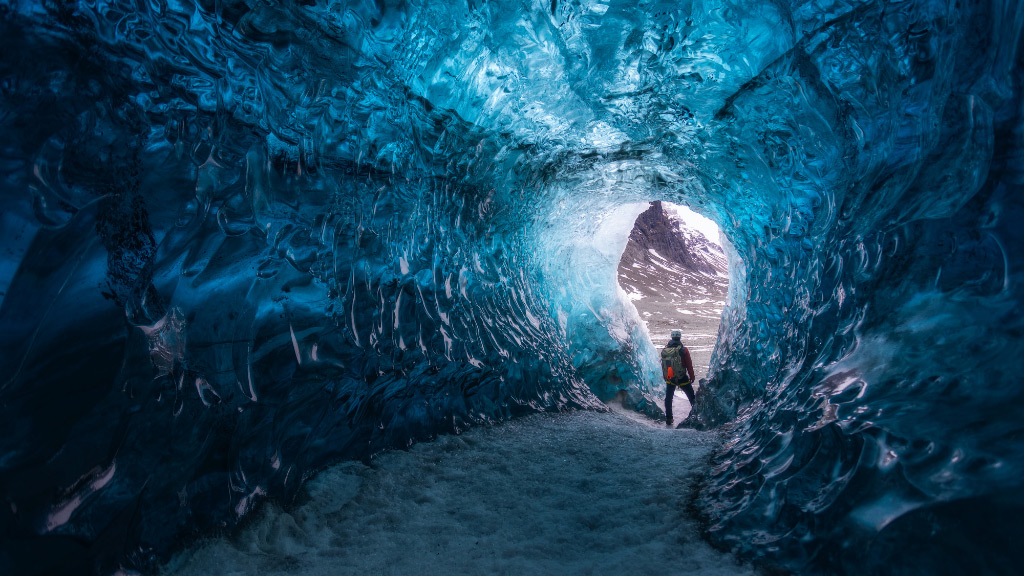 Why-Iceland’s-Ice-Caves-are-Changing_-the-Impact-of-Melting-Glaciers---Pic-4 A person standing in the mouth of an ice cave looking out with blue ice all around him.